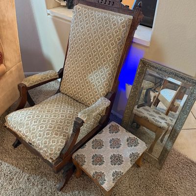 Antique carved wooden platform rocking chair with patterned upholstery, paired with a matching small footstool and an ornate framed wall...