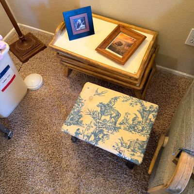 Small upholstered footstool featuring a blue toile pattern, placed next to a set of wooden nesting TV tray tables with framed artwork.
