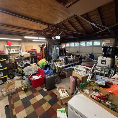 Crowded garage filled with assorted household items, including storage totes, luggage, hand tools, rolling carts, and clothing racks.