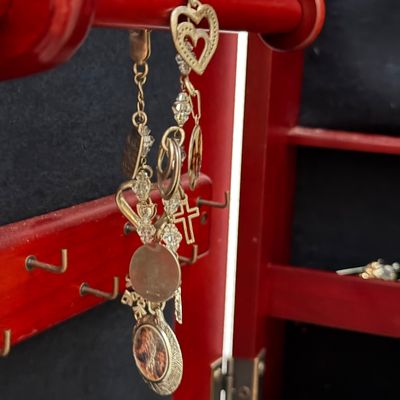 Gold-tone charm bracelet featuring hearts, a cross, and beaded accents, hanging inside a wooden jewelry armoire.