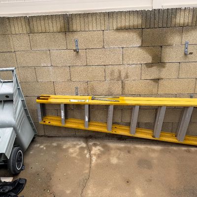 Yellow fiberglass extension ladder shown resting horizontally against a concrete block wall next to a plastic lawn cart.