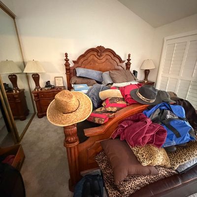 Traditional carved wood king bed frame with matching nightstands and lamps, surrounded by various hats, textiles, and bags.