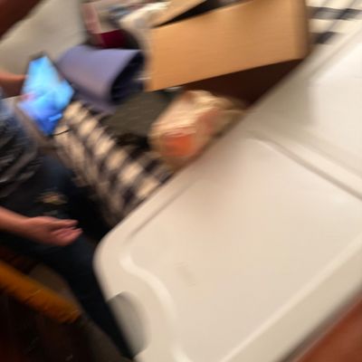 A blurry view of various storage items, including a white plastic container, cardboard boxes, and bedding on a bed.