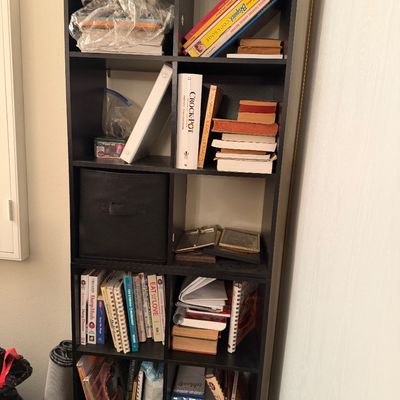 Tall black laminate cube bookcase filled with assorted cookbooks, notebooks, binders, and small household items.