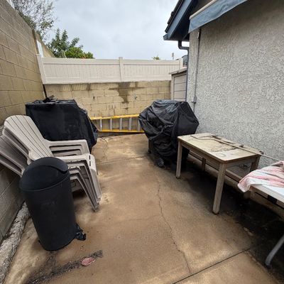 Outdoor patio area featuring stacked plastic lawn chairs, a covered barbecue grill, a trash receptacle, and a weathered utility table.