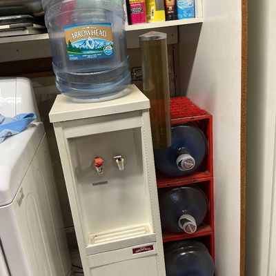 Sale Photo Thumbnail #131: Freestanding Arrowhead water dispenser with a top-mounted 5-gallon jug, side cup holder, and a stack of three additional water jugs stored in red plastic crates.