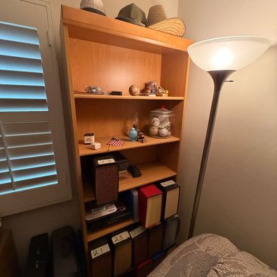 Sale Photo Thumbnail #103: Tall light wood finish open bookcase filled with small decor, collectibles, and magazine organizers, alongside a modern metal floor lamp.