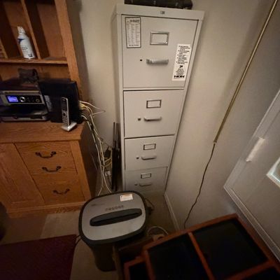 Sale Photo Thumbnail #102: Tall grey HON four-drawer metal filing cabinet positioned alongside a wooden desk unit and paper shredder.