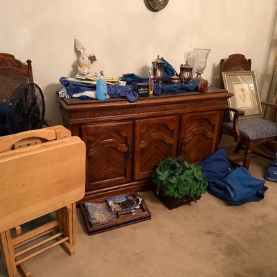 Traditional oak buffet cabinet surrounded by wooden TV tray tables, an Oreck upright vacuum, and high-back dining chairs.
