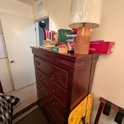 Traditional dark wood highboy dresser featuring ornate brass handles, topped with various books and a woven table lamp.