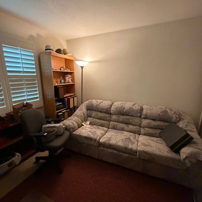 Home office setup featuring a patterned three-cushion sofa, a rolling desk chair, a light wood bookshelf, and a floor lamp.