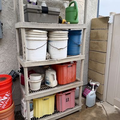 Four-tier beige plastic storage shelving unit holding assorted gardening supplies, buckets, and plastic crates.