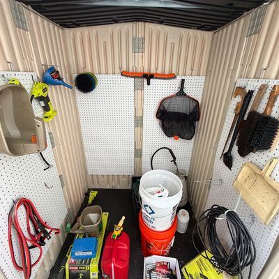 Plastic outdoor storage shed filled with various lawn and garden tools, buckets, jumper cables, and Ryobi power equipment.