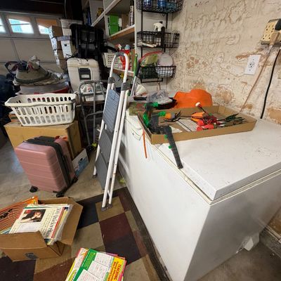 White chest freezer surrounded by miscellaneous household items including a pink rolling suitcase, folding step ladder, and boxes of...