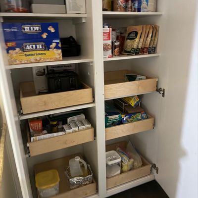 Tall white wooden kitchen pantry cabinet featuring dual doors and pull-out wooden drawers filled with assorted food items and small...