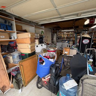 Crowded garage packed with various household goods, including a bicycle, large water jugs, luggage, folding tables, and stacked storage...