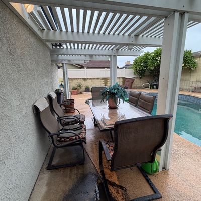 Outdoor patio dining set under a pergola, featuring a tile-top rectangular table and matching metal spring-motion chairs with mesh fabric...