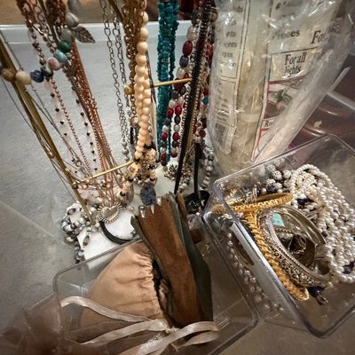 Collection of fashion jewelry including beaded necklaces on a brass display stand, faux pearl strands, and metal bangles in a clear...