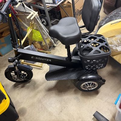 Black 3-wheeled seated electric scooter featuring a wide foot deck, padded seat with backrest, and rear storage basket.