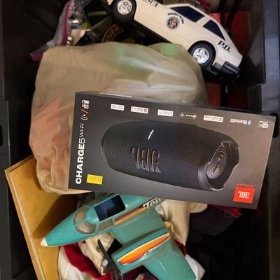 Black plastic storage tub filled with various items, including a boxed JBL Charge 5 Wi-Fi portable speaker, a vintage turquoise Tonka...