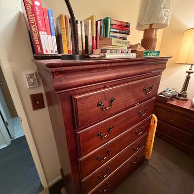Traditional dark cherry wood tallboy chest of drawers with bail handles, topped with an assortment of books and a desk lamp.