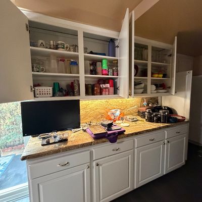 Open kitchen cabinets filled with various drinkware, mugs, and plastic cups, alongside a granite countertop featuring a computer monitor...