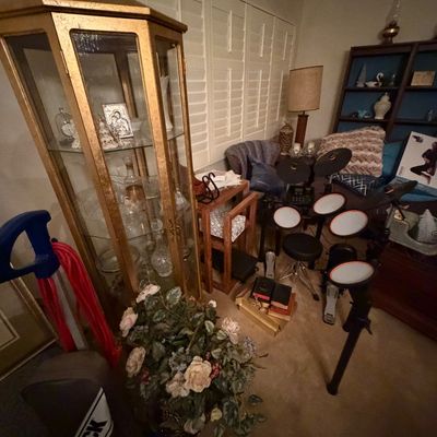 Gold-toned hexagonal curio cabinet displayed alongside an electronic drum kit, Oreck vacuum cleaner, and a floral arrangement.