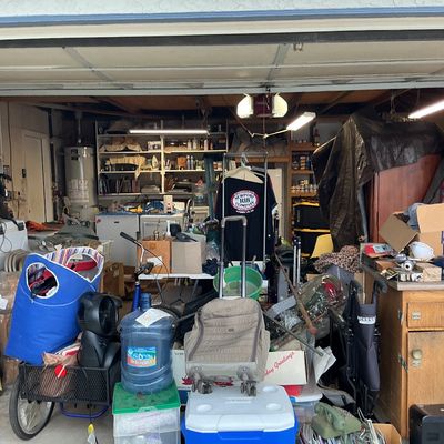 Sale Photo Thumbnail #127: A crowded garage filled with diverse items including a blue utility cart, rolling luggage, plastic storage tubs, tools, and wooden furniture.