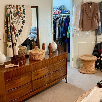 Sale Photo Thumbnail #11: Mid-century modern walnut triple dresser with matching mirror a pair of white ceramic table lamps, woven baskets, vintage clothing hanging on the door, and a woven hourglass stool
