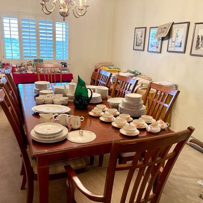 Sale Photo Thumbnail #6: Large wooden dining table set with Vernon Ware, Pfalzgraff, and mid-century modern china, green Blenko glass pitcher, and surrounding slat-back chairs in a dining room with framed art