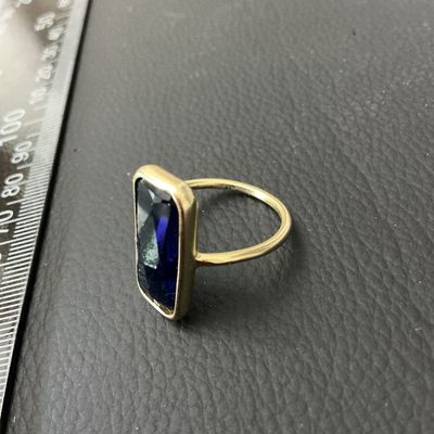 Sale Photo Thumbnail #231: A gold-tone ring featuring a large bezel-set emerald-cut faceted blue stone, likely glass or synthetic crystal. The ring band is thin and round in profile. Shown on a mandrel measuring approximately US size 8 (EU size 57/58). Light general wear to metal f