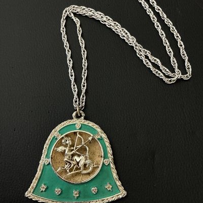 Sale Photo Thumbnail #196: A vintage silver-tone rope chain necklace featuring a bell-shaped pendant with a green enamel border and a central round medallion depicting the Sagittarius archer/cherub with a bow and arrow. Pendant measures approximately 60mm wide by 60mm tall. Shows l