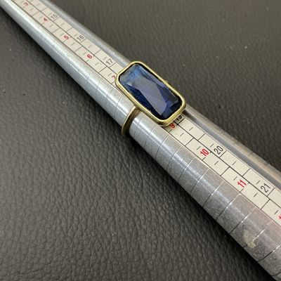 Sale Photo Thumbnail #226: A gold-tone ring featuring a large bezel-set emerald-cut faceted blue stone, likely glass or synthetic crystal. The ring band is thin and round in profile. Shown on a mandrel measuring approximately US size 8 (EU size 57/58). Light general wear to metal f