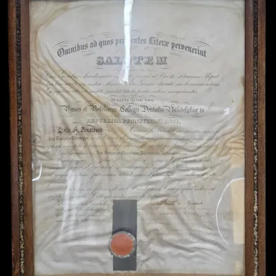 Sale Photo Thumbnail #49: Framed 1868 dental diploma from the Philadelphia Dental College, featuring a red wax seal and set in a carved wooden frame showing significant age and creasing.