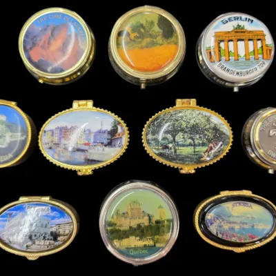 Sale Photo Thumbnail #72: Collection of nine vintage souvenir pill boxes featuring scenic European and Canadian landmarks, including Berlin, Québec, and Stresa.