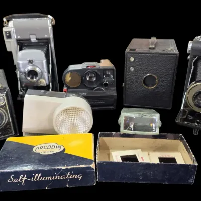 Sale Photo Thumbnail #100: Collection of vintage cameras and accessories, including Kodak folding cameras, box cameras, a Polaroid Land camera, and an Arcadia self-illuminating slide viewer with original box.
