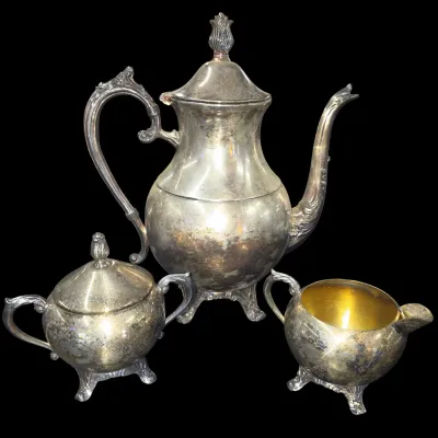 Sale Photo Thumbnail #147: Three-piece vintage silver-plated tea service, including a tall teapot, covered sugar bowl, and gold-washed cream pitcher, all featuring ornate handles and finials with surface tarnish.