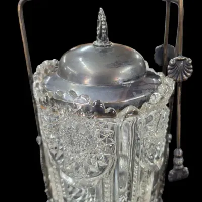 Sale Photo Thumbnail #83: Victorian-style silver-plate pickle castor set with a pattern-cut glass jar, matching lid, hanging tongs, and ornate scrollwork handle.