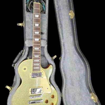 Sale Photo Thumbnail #29: Epiphone Les Paul electric guitar in a gold flake finish, displayed inside its gray plush-lined hard shell case.