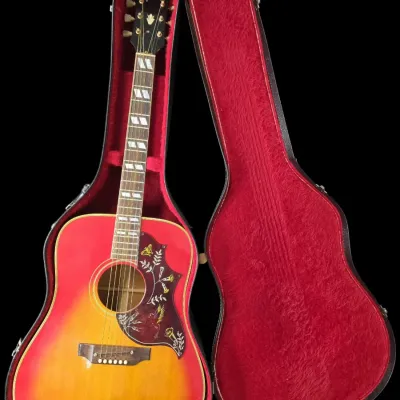 Sale Photo Thumbnail #24: Gibson Hummingbird acoustic guitar with a cherry sunburst finish and ornate engraved pickguard, resting in a red plush-lined hard shell case.