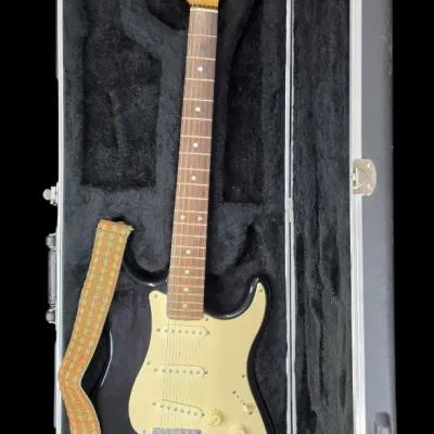 Sale Photo Thumbnail #31: Black Stratocaster-style electric guitar with a vintage patterned strap, housed inside a plush-lined hard shell case.