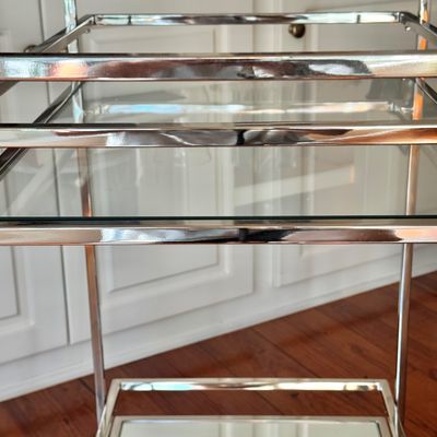 Sale Photo Thumbnail #272: A sleek, minimalist bar cart characterized by a polished chrome-plated metal frame and dual -tier shelving. The upper tier is a clear glass surface, while the lower tier is fitted with a mirrored glass shelf, providing a sophisticated reflected quality. T