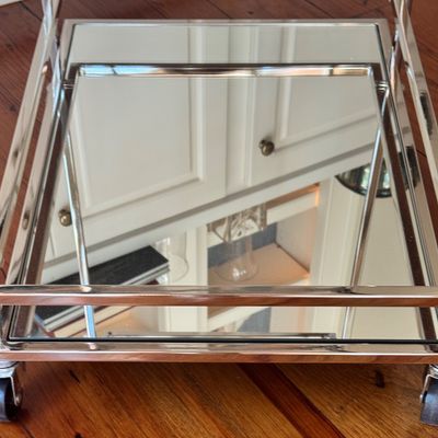 Sale Photo Thumbnail #271: A sleek, minimalist bar cart characterized by a polished chrome-plated metal frame and dual -tier shelving. The upper tier is a clear glass surface, while the lower tier is fitted with a mirrored glass shelf, providing a sophisticated reflected quality. T