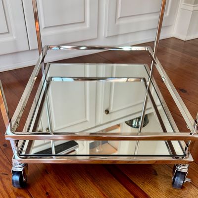 Sale Photo Thumbnail #273: A sleek, minimalist bar cart characterized by a polished chrome-plated metal frame and dual -tier shelving. The upper tier is a clear glass surface, while the lower tier is fitted with a mirrored glass shelf, providing a sophisticated reflected quality. T