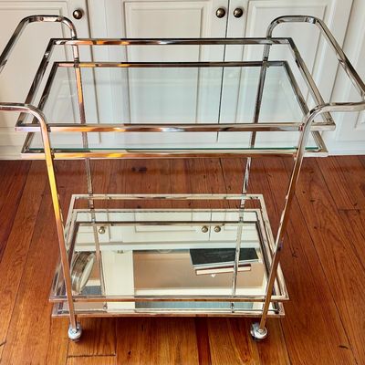 Sale Photo Thumbnail #268: A sleek, minimalist bar cart characterized by a polished chrome-plated metal frame and dual -tier shelving. The upper tier is a clear glass surface, while the lower tier is fitted with a mirrored glass shelf, providing a sophisticated reflected quality. T