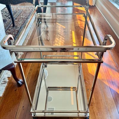 Sale Photo Thumbnail #270: A sleek, minimalist bar cart characterized by a polished chrome-plated metal frame and dual -tier shelving. The upper tier is a clear glass surface, while the lower tier is fitted with a mirrored glass shelf, providing a sophisticated reflected quality. T