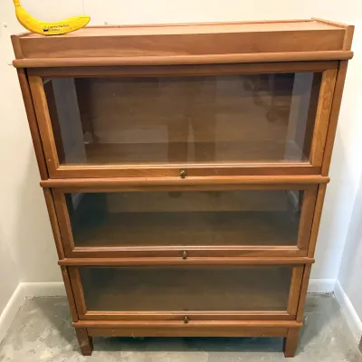 Sale Photo Thumbnail #10: Three-shelf barrister bookcase made of light finish wood featuring glass lift-up doors with small brass knobs.