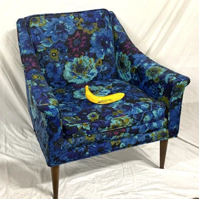 Sale Photo Thumbnail #9: Vintage mid-century modern armchair featuring bold blue and green floral jacquard upholstery and tapered wooden legs. A yellow banana prop sits on the seat cushion for scale.