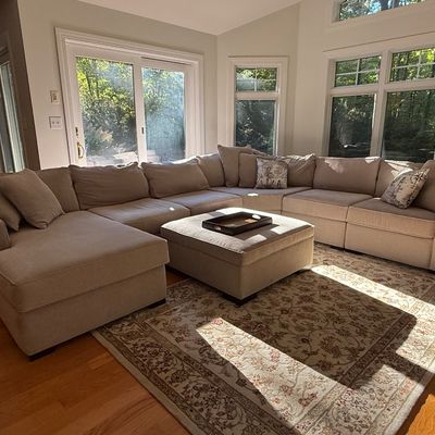 Sale Photo Thumbnail #2: Large contemporary neutral-upholstered sectional sofa with matching square ottoman coffee table, set over an oriental-style area rug in a sunlit room.