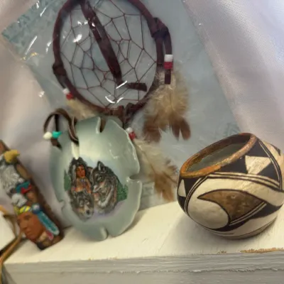 Sale Photo Thumbnail #40: Collection of Native American style souvenir crafts, including a packaged feather dreamcatcher, a painted ceramic sand dollar pendant with wolf artwork, a carved wooden totem piece, and a small pottery vessel with geometric designs.